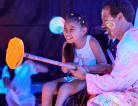 A smiling little girl is sitting in her wheelchair. Dwayne is helping her hold a paint pole and they are getting ready to paint on a vertical surface. There are other people in the background.