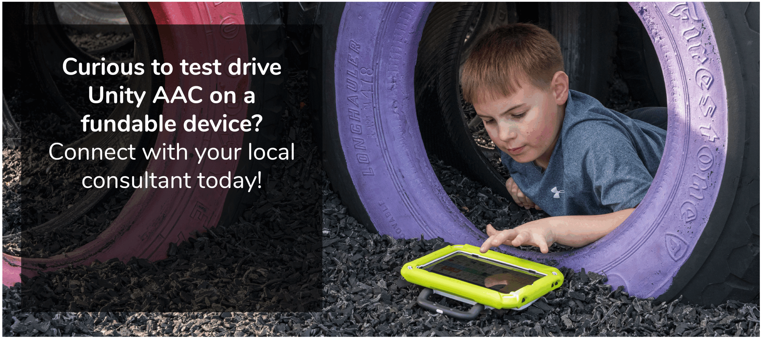A boy laying on his belly with a tire around him. The boy is pushing a button on a Unity AAC device on the ground